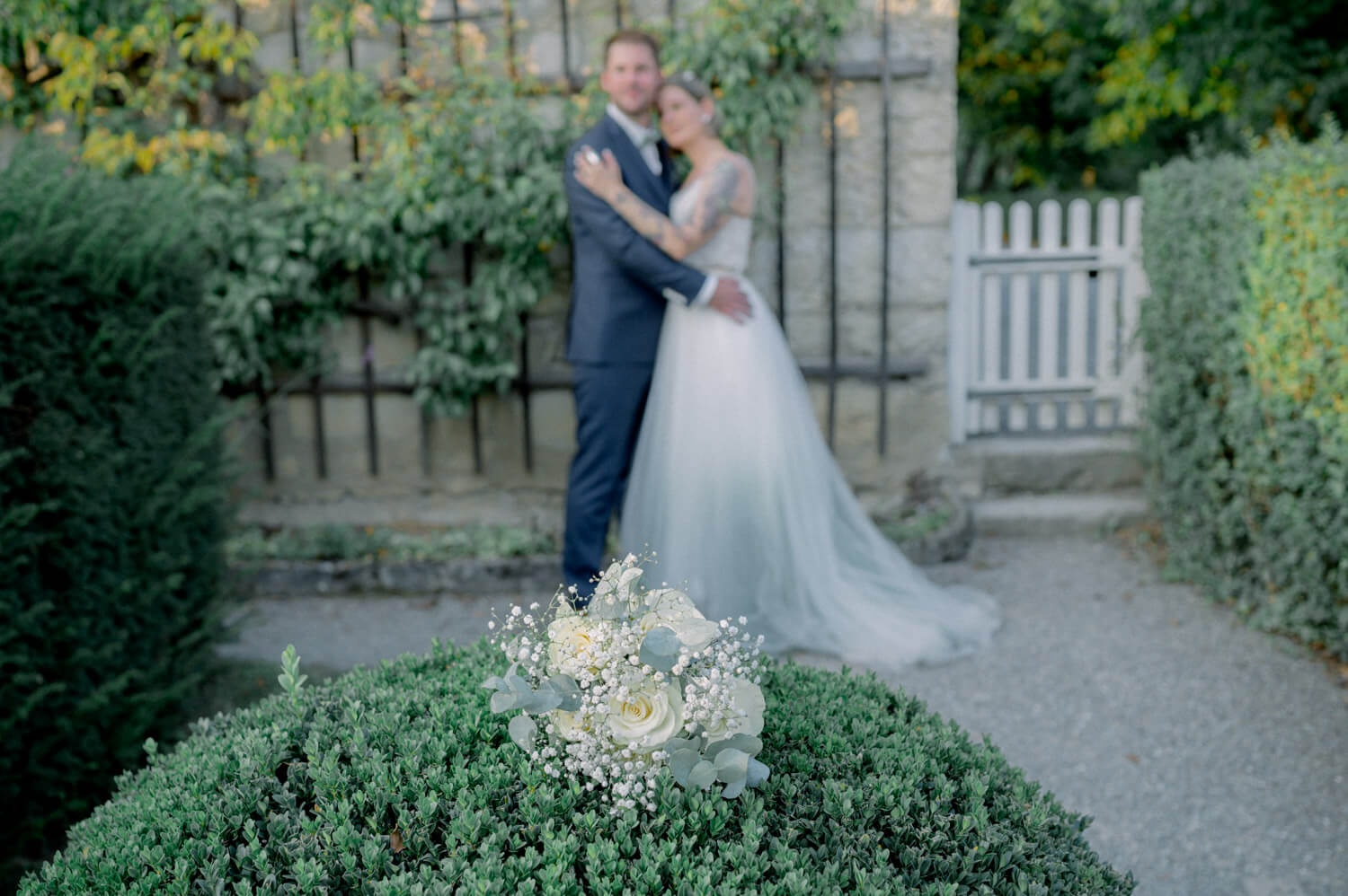 Hochzeitsblumen liegen im Vordergrund, das Brautpaar steht unscharf im Hintergrund im Garten