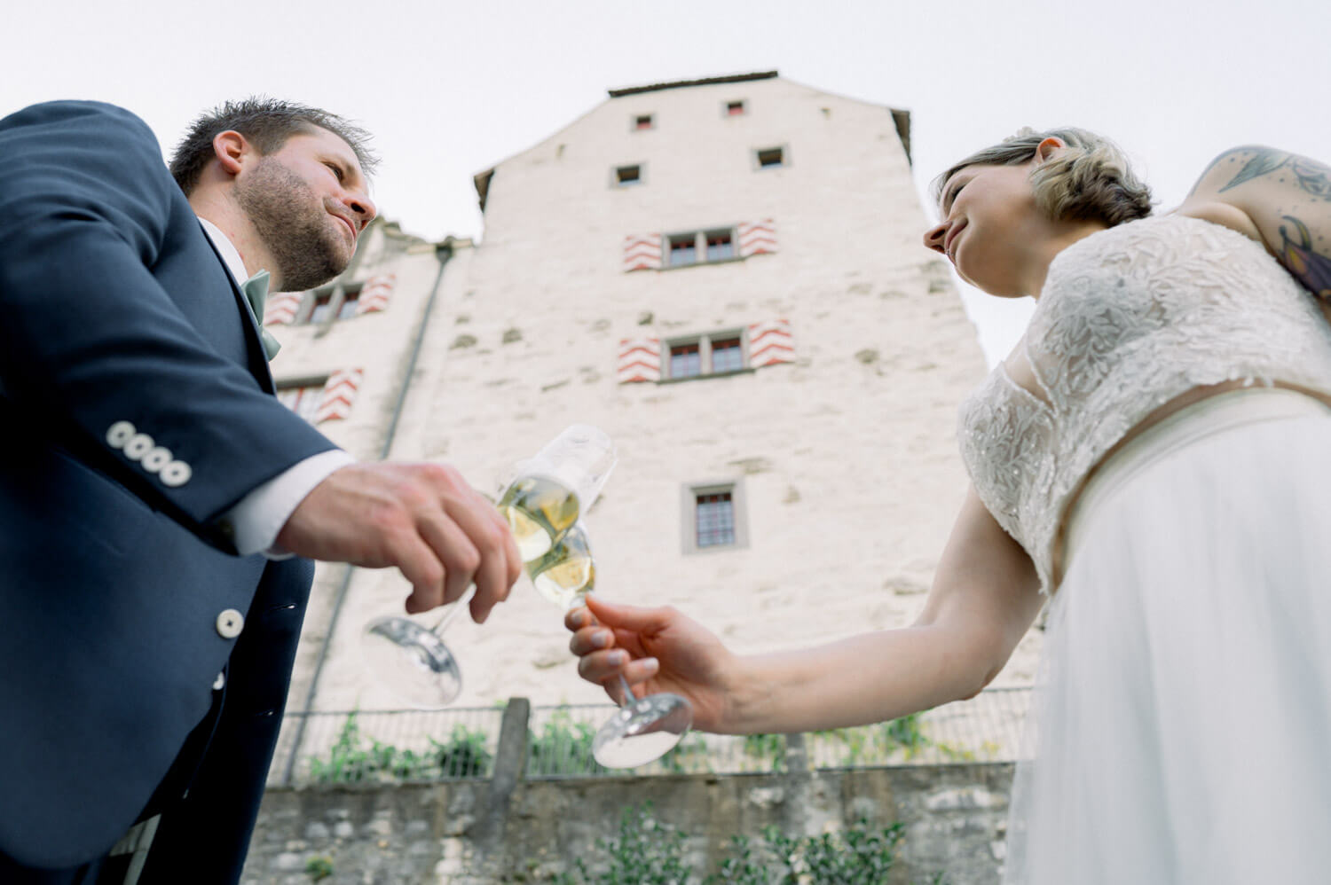 Braut und Bräutigam stossen mit Champagnergläsern vor einem historischen Schloss an, fotografiert aus der Untersicht.