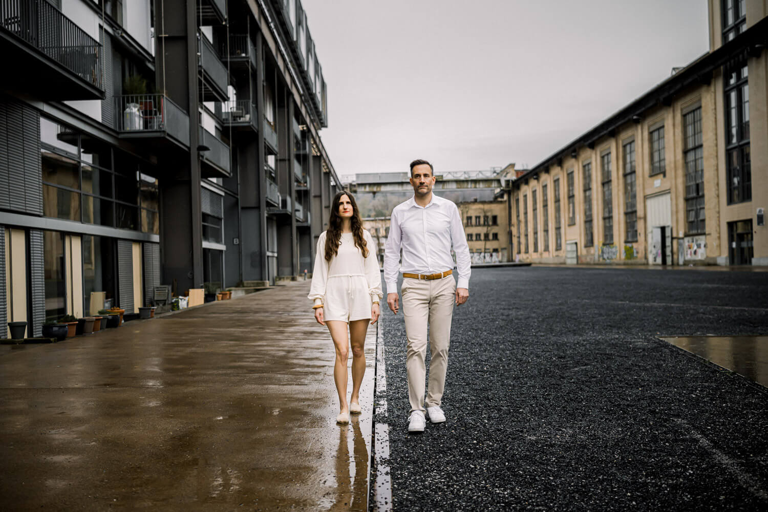 Paar geht frontal durch urbanes Industrieareal mit klaren Linien und ruhiger Stimmung