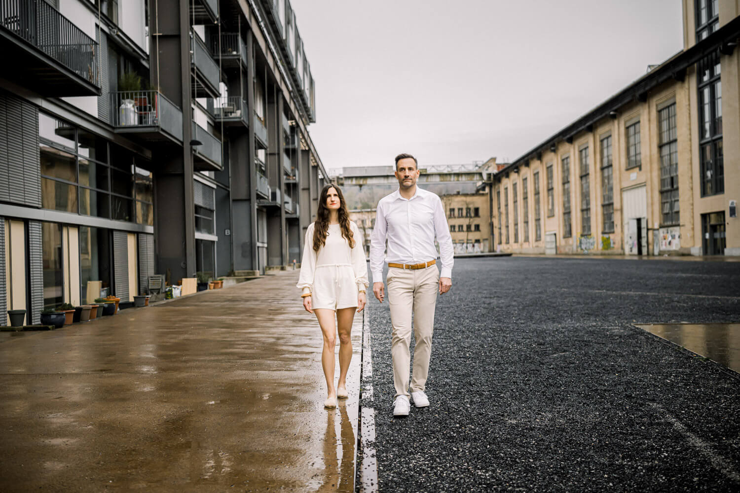 Paar geht frontal durch urbanes Industrieareal mit klaren Linien und ruhiger Stimmung